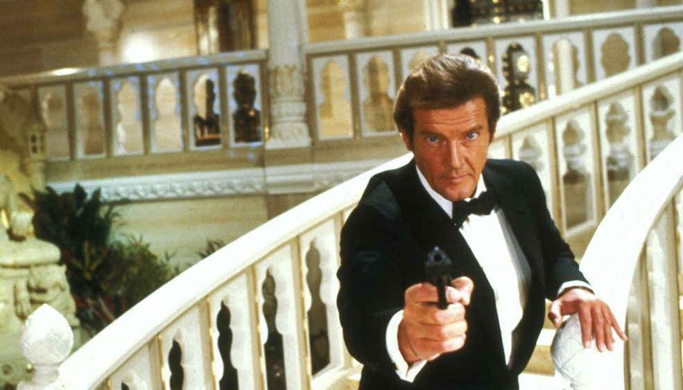 Roger Moore jako James Bond ve smokingu míří pistolí na točitém mramorovém schodišti