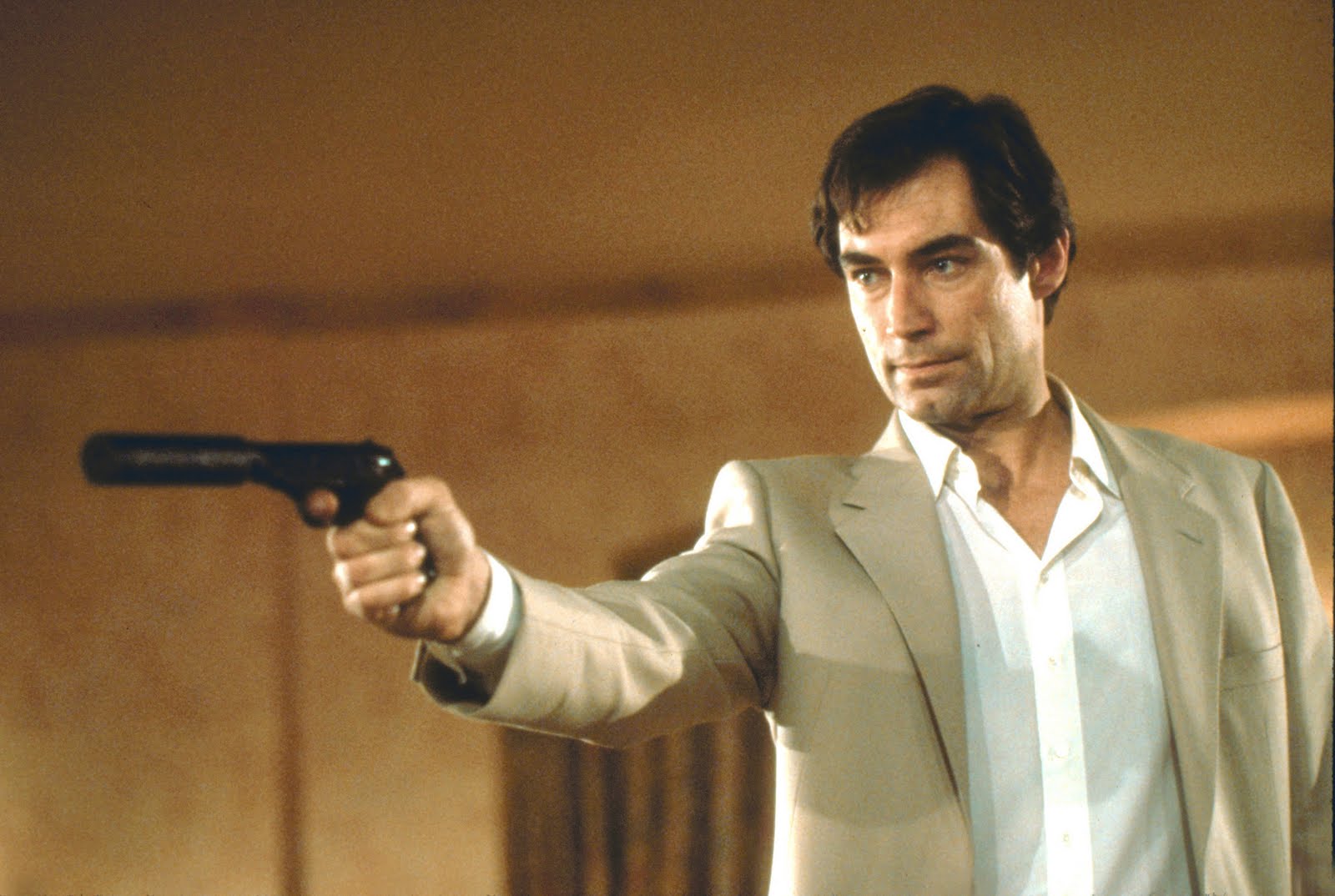Timothy Dalton jako James Bond v krémovém obleku míří pistolí s tlumičem