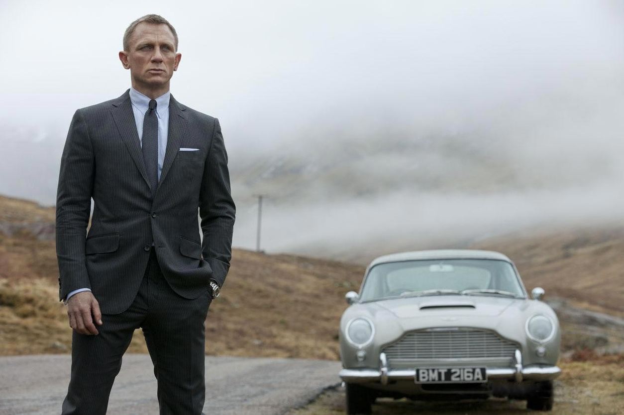 Daniel Craig jako James Bond v obleku stojí na silnici vedle stříbrného Aston Martinu DB5 se značkou BMT 216A