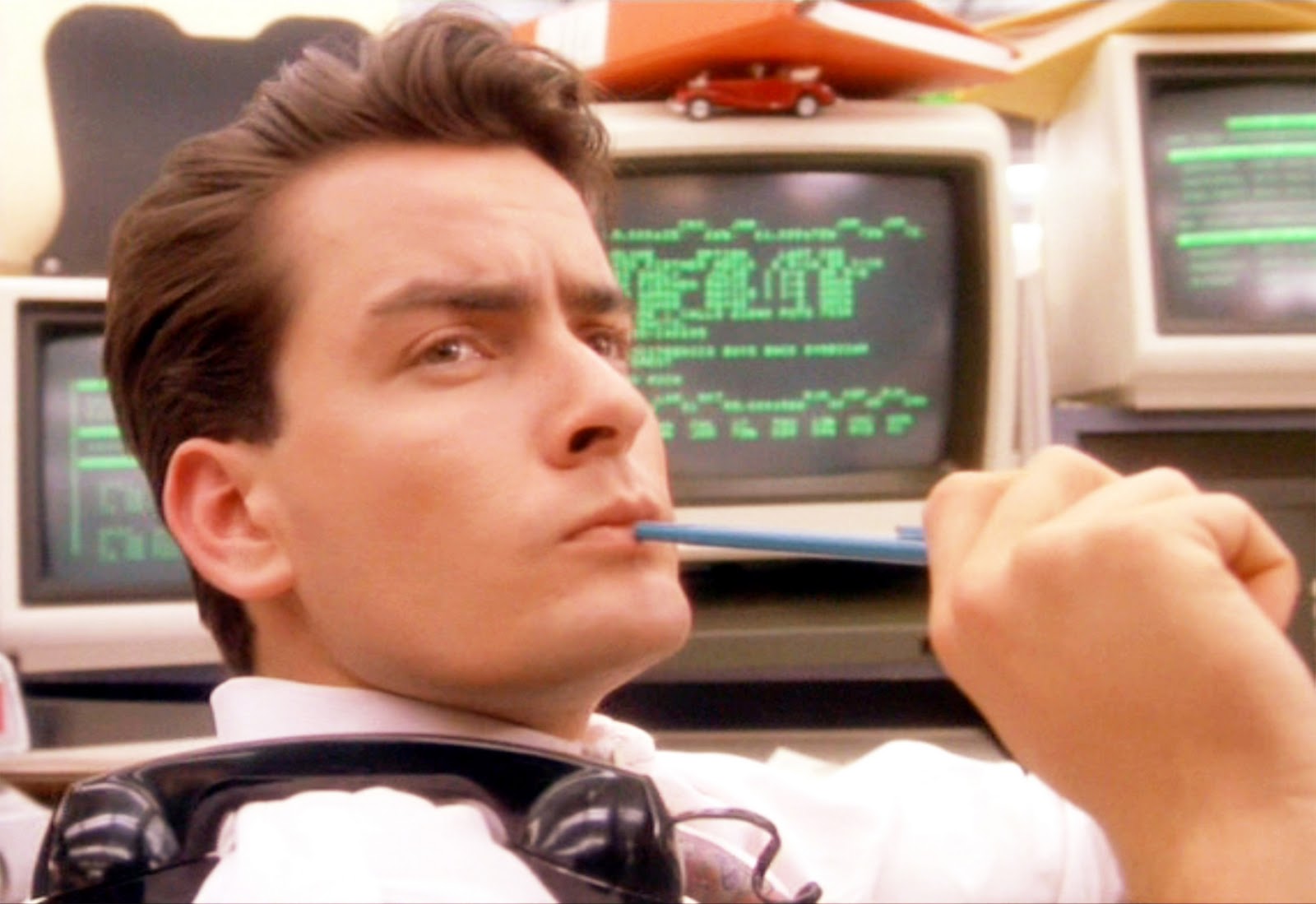 Charlie Sheen jako Bud Fox ve filmu Wall Street, s tužkou v ústech před obrazovkami s burzovními kurzy