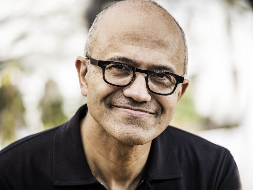 Muž s brýlemi a plnovousem v tmavém polo tričku se usmívá do kamery - šéf Microsoftu Satya Nadella