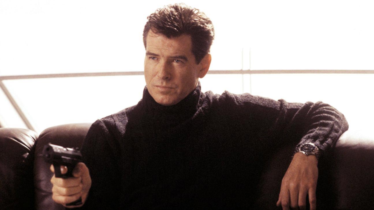 Elegantní herec Pierce Brosnan jako James Bond v černém svetru, pistolí a luxusními hodinkami.