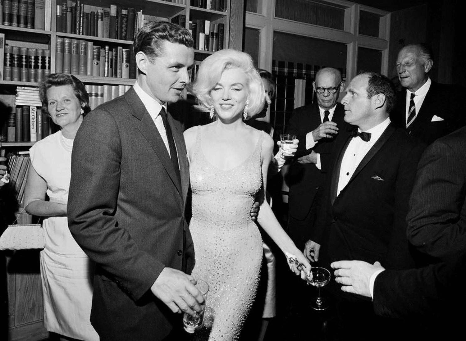 Černobílá fotografie Marilyn Monroe v třpytivých šatech na společenské akci obklopené muži ve smokingu