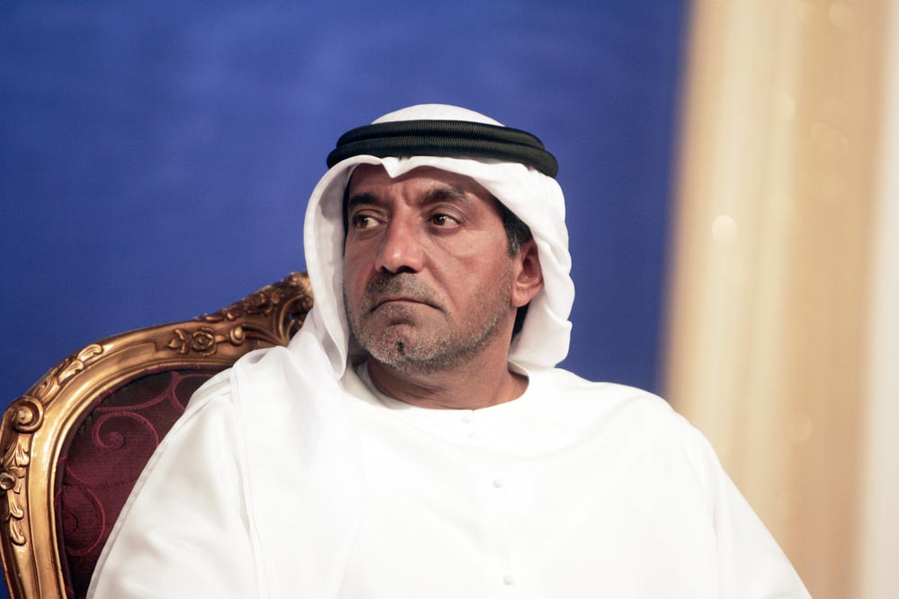 Muž v bílém tradičním arabském oděvu s černou agalou sedící ve zlacené židli s červeným čalouněním