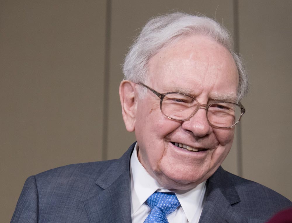 Warren Buffett usmívající se v brýlích, šedém obleku a modré kravatě