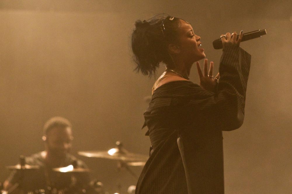 Zpěvačka Rihanna zpívá do mikrofonu na koncertě, v pozadí bubeník