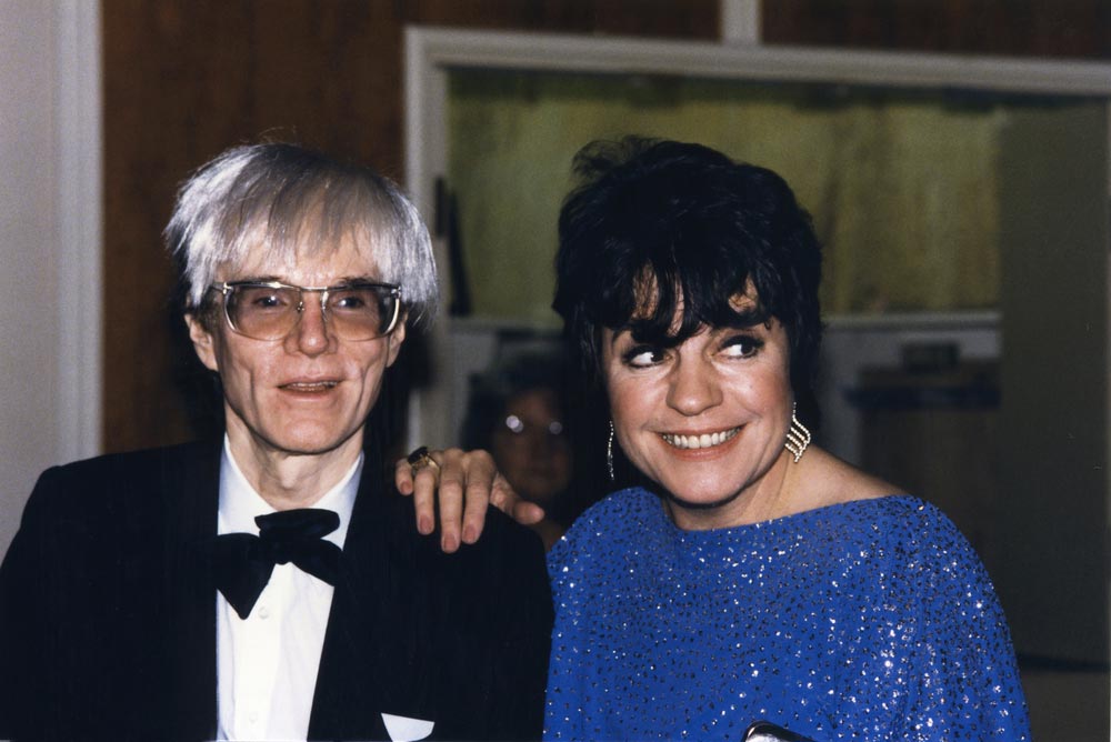 Andy Warhol s typickou stříbrnou parukou a brýlemi po boku ženy v modrých třpytivých šatech