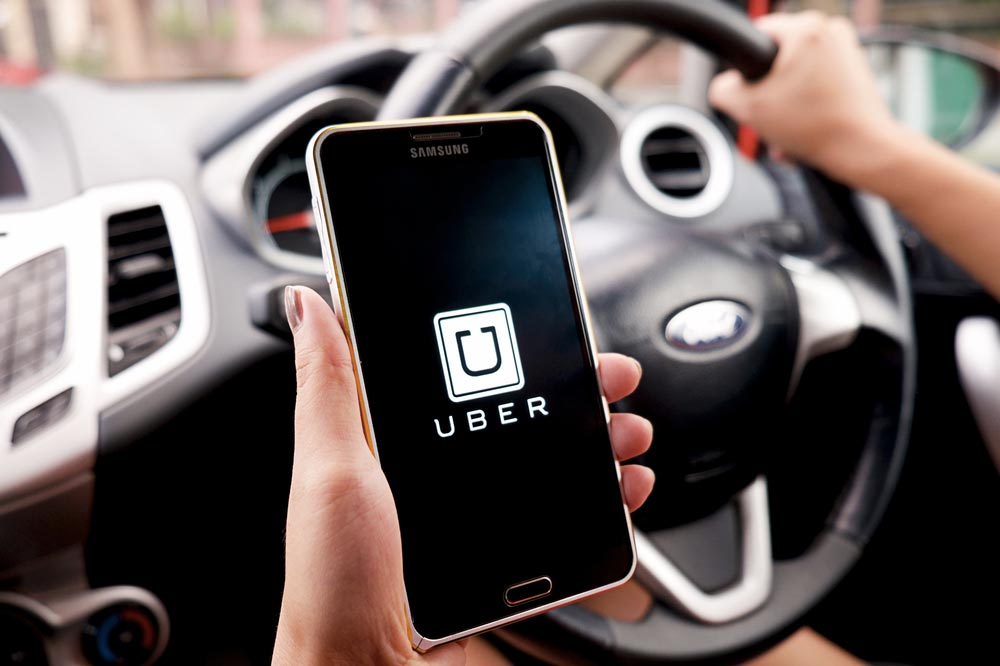 Ruka držící telefon Samsung se svíceným logem Uber, v pozadí volant a ruka řidiče