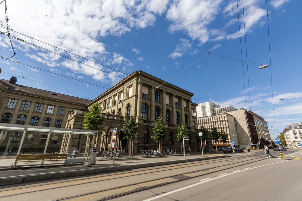 Historizující budova univerzity s tramvajovými kolejemi a zastávkou před vstupem, cyklisté opřeni o zábradlí