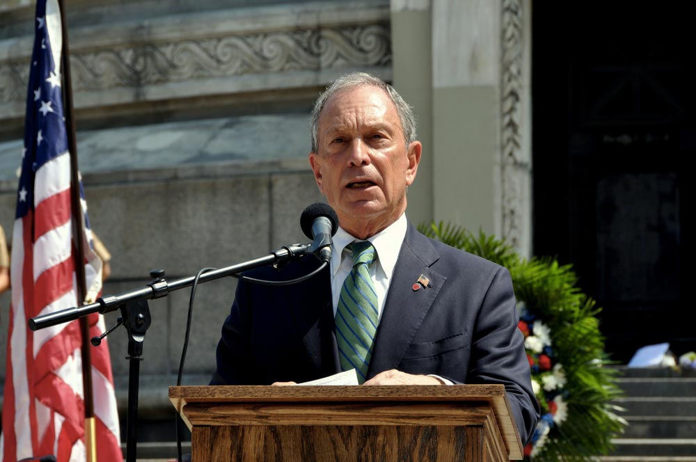Michael Bloomberg s mikrofonem u řečnického pultu, vedle americká vlajka