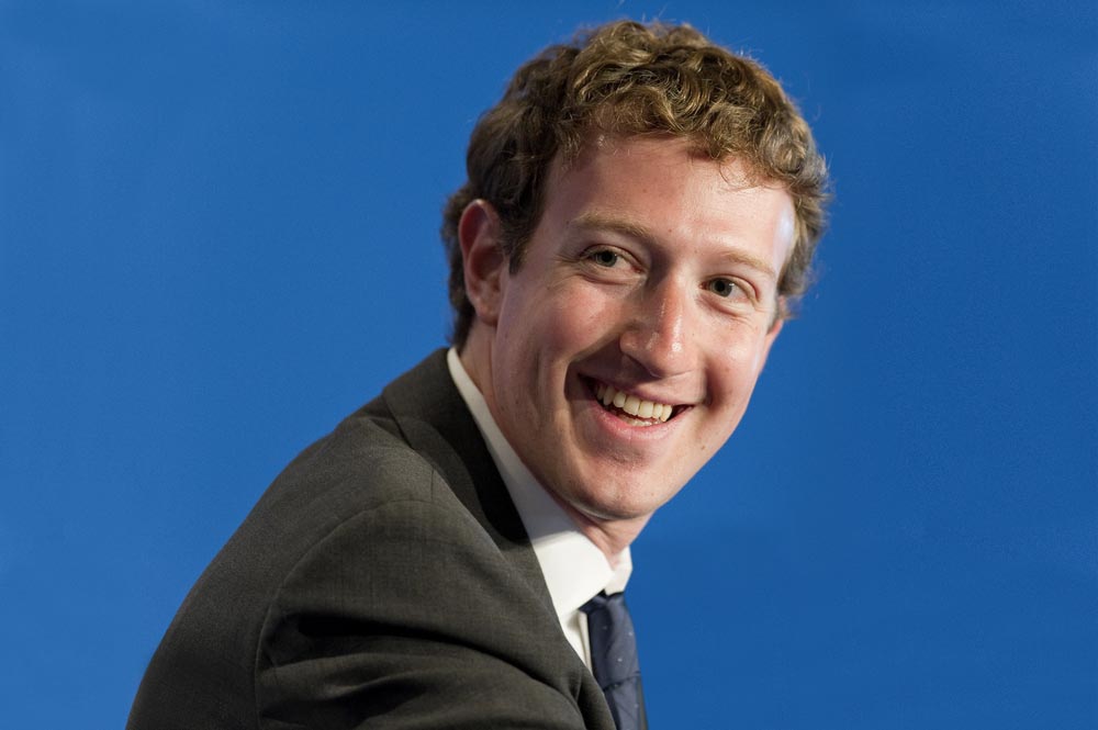 Mark Zuckerberg v tmavém obleku s kravatou, usmívající se na modrém pozadí