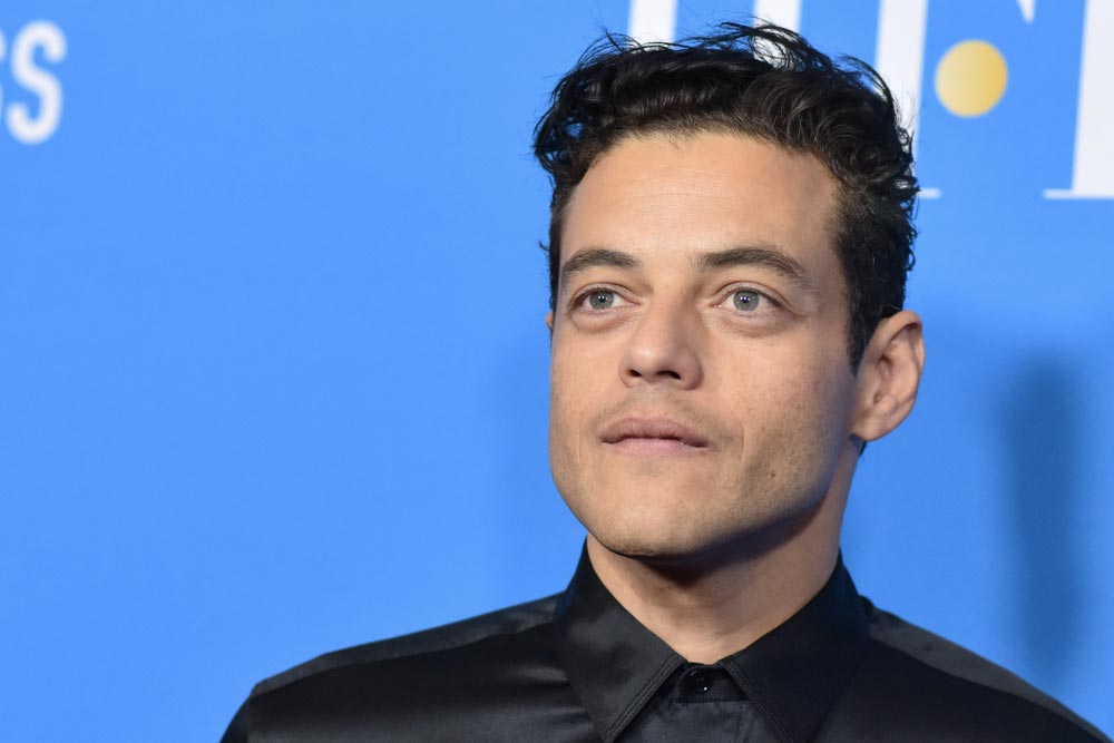 Rami Malek s tmavými vlnitými vlasy a černou saténovou košilí, portrét na modrém pozadí