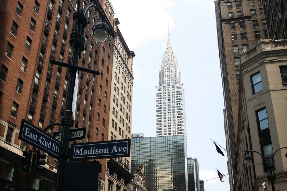 Mrakodrap Chrysler Building s věží ve stylu art deco mezi okolními budovami, v popředí uliční cedule East 42nd St a Madison Ave