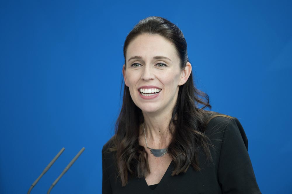 Jacinda Ardern s tmavými vlasy usměvavě mluví na modrém pozadí, v popředí mikrofony
