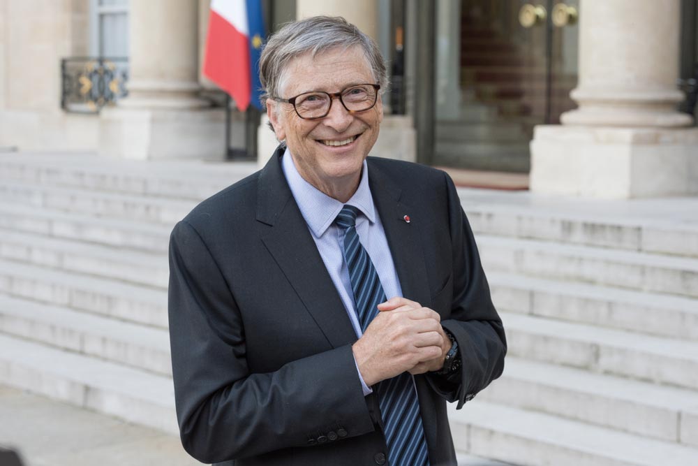 Bill Gates v tmavém obleku a pruhované kravatě před budovou s francouzskou vlajkou
