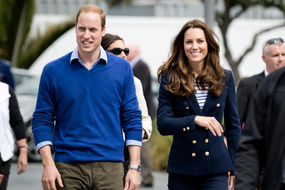 Princ William v modrém svetru vedle usmívající se Kate Middleton, v pozadí ochranka
