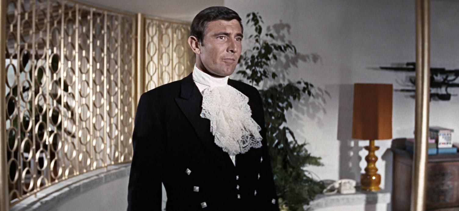 George Lazenby jako James Bond v černém fraku s krajkovým jabotem stojí v interieru se zlatou dekorativní mřížkou