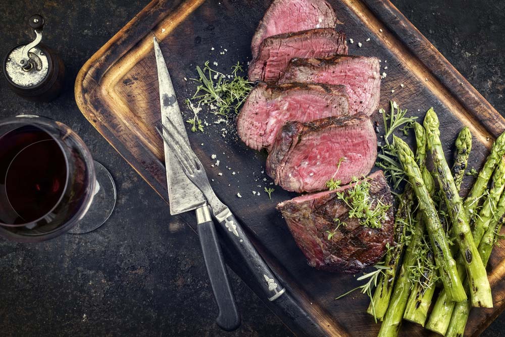 Nakrájený steak medium rare s chřestem a bylinkami na dřevěném prkénku, vedle sklenka červeného vína