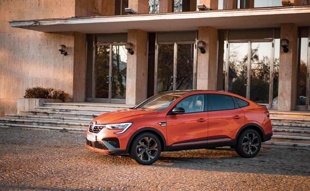 Oranžové SUV Renault Arkana zaparkované před budovou se skleněnými dveřmi a sloupy
