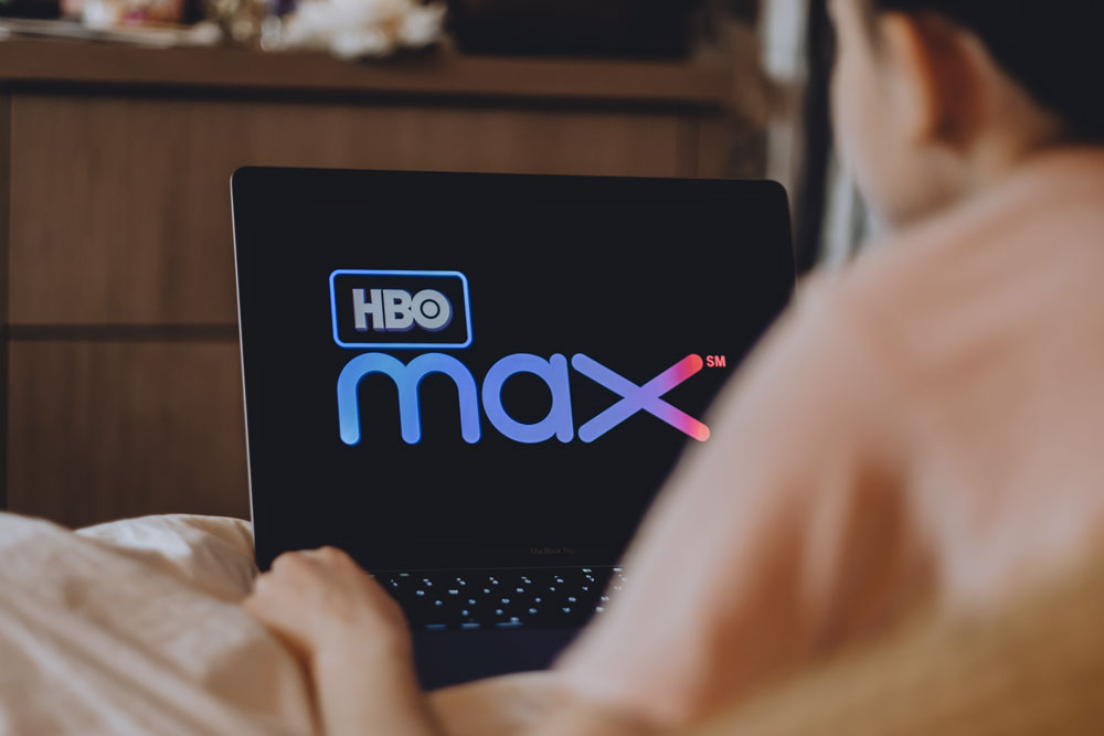 Notebook umístěný na posteli v ložnici, na jehož obrazovce je zobrazeno logo služby HBO Max.